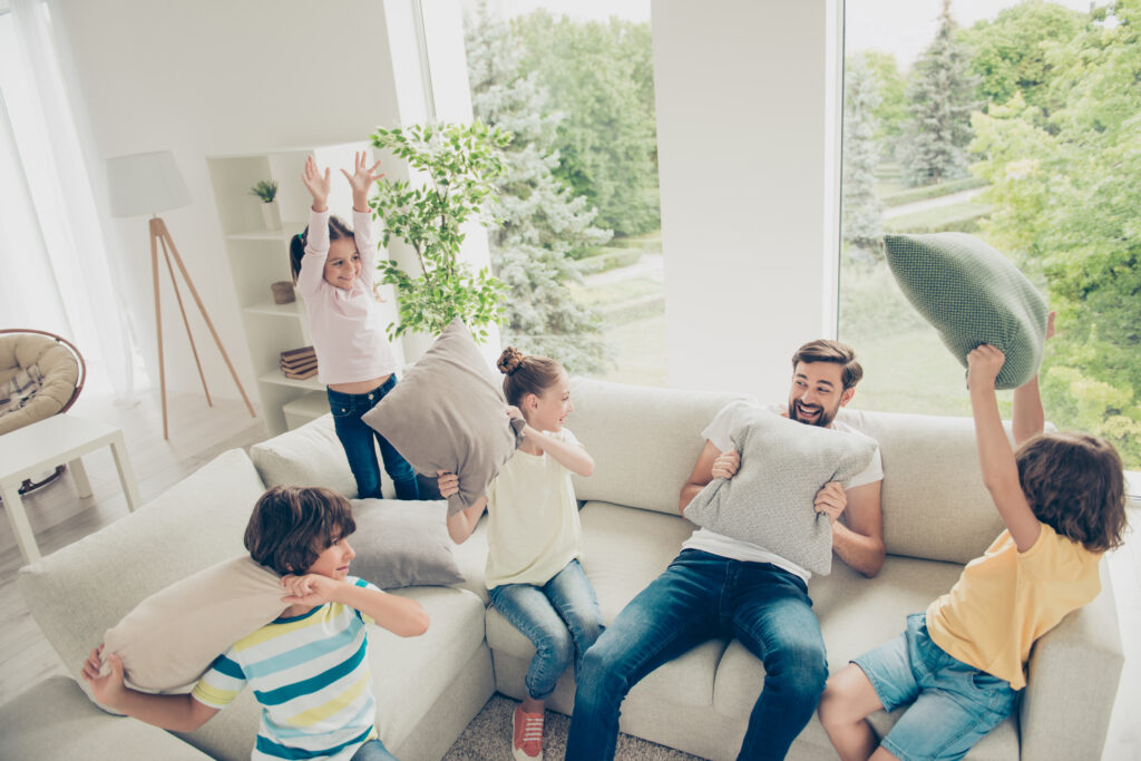 Sala com família reunida, pai e quatro crianças sentados em sofá em L, segurando almofadas e com expressões divertidas, sugerindo um momento de brincadeira e descontração.